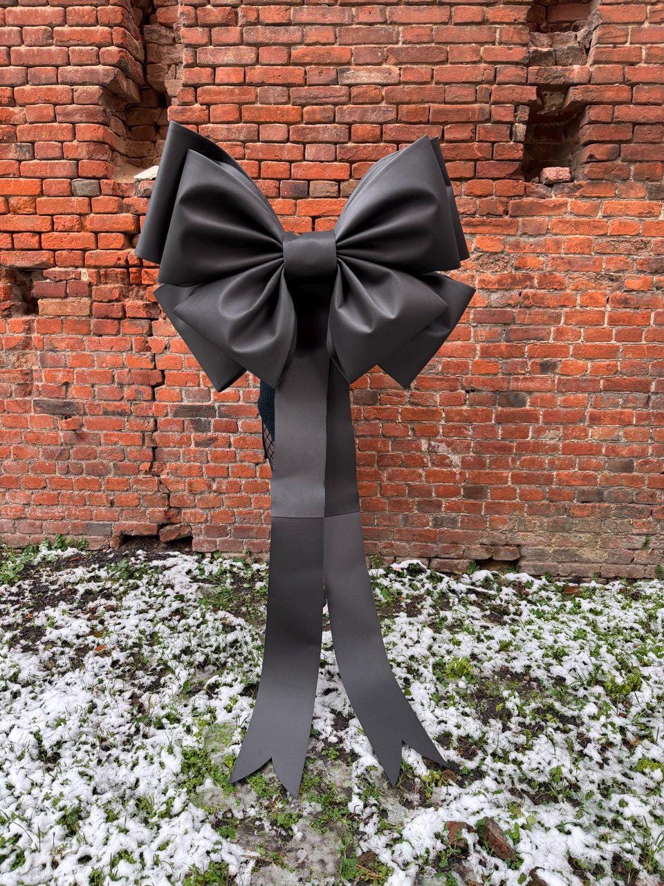 May include: A large, black decorative bow with long, flowing tails. The bow is made of a matte material and is positioned in front of a weathered brick wall. The ground is covered with patches of snow and green grass.