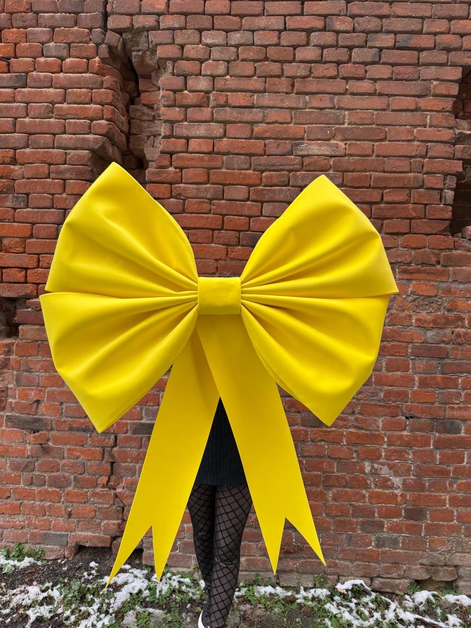May include: A large, bright yellow bow with long, pointed streamers. The bow is made of a smooth, possibly satin-like material. The background is a weathered brick wall.