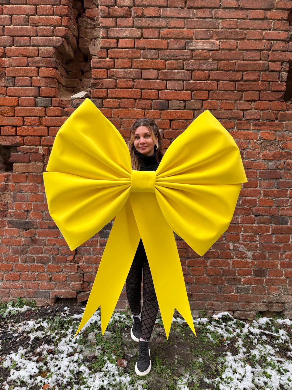 May include: A large, bright yellow bow with long streamers. The bow is the focal point, set against a weathered brick wall. The bow's size suggests it could be used for decoration or as a prop.