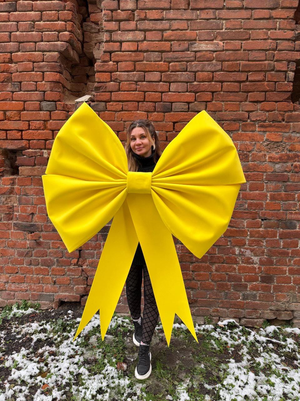 May include: A large, bright yellow bow with long, pointed streamers. The bow is the focal point, set against a weathered brick wall. The bow's size suggests it could be used for decoration or as a prop.