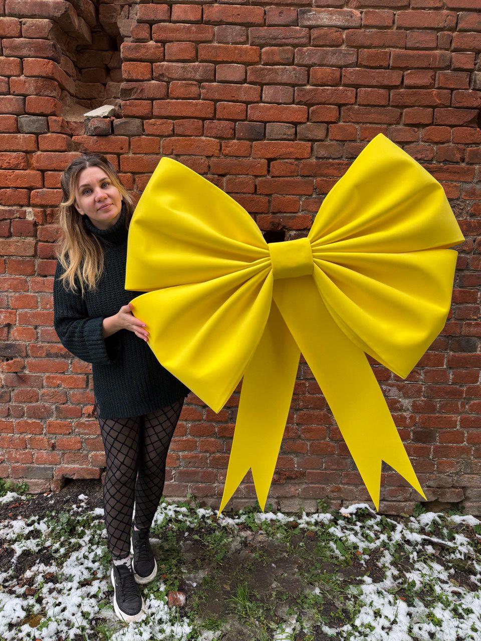 May include: A large, bright yellow bow, possibly for decoration. The bow is made of a smooth material and has long, flowing ribbons. It is positioned against a red brick wall.