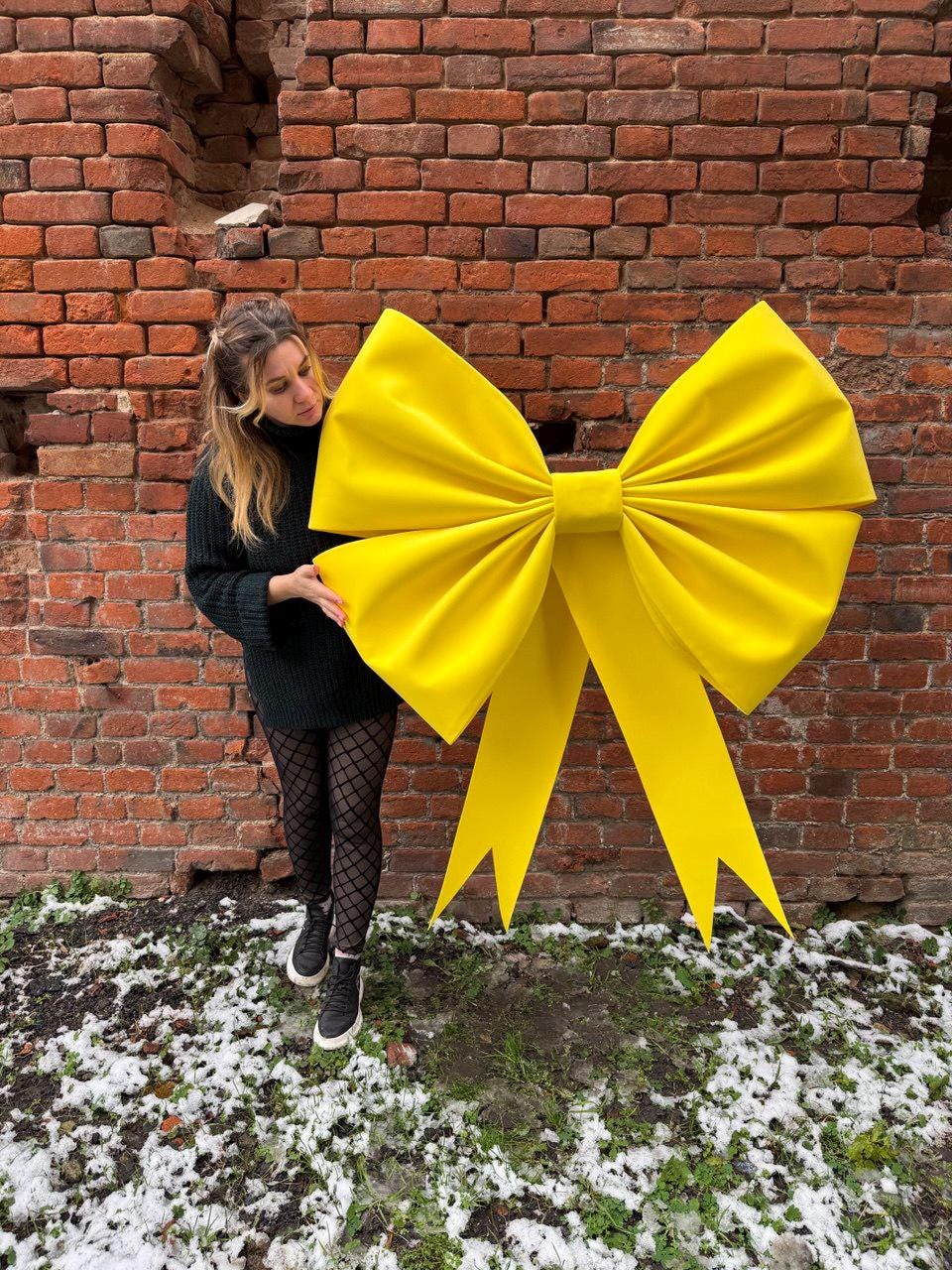 May include: A large, bright yellow bow against a red brick wall. The bow has a classic shape with two long, pointed tails. The bow is a vibrant, eye-catching decoration.