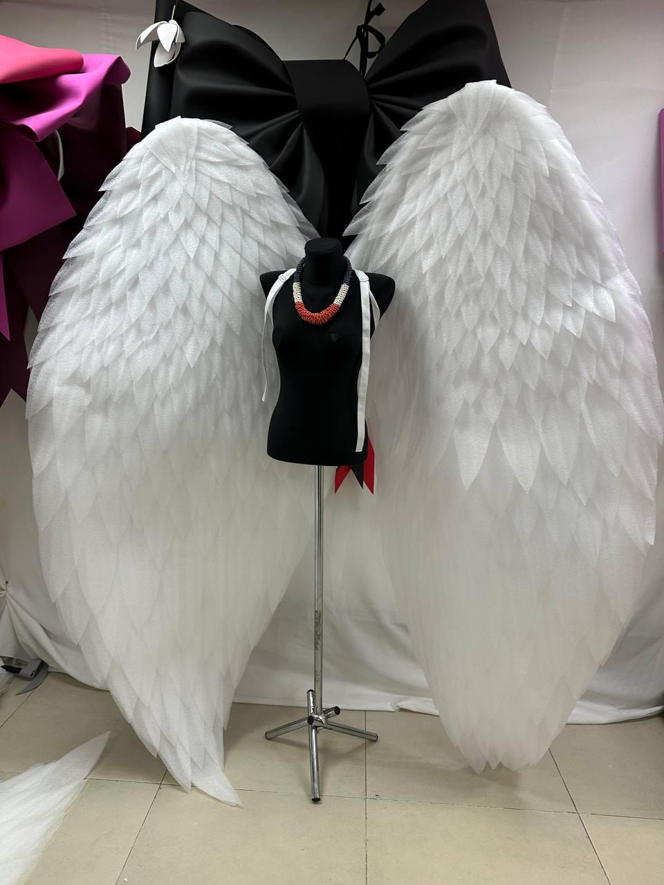 May include: Large white angel wings with a layered feather design are displayed behind a black mannequin. The mannequin wears a black top and a red and white beaded necklace. A large black bow is at the top.