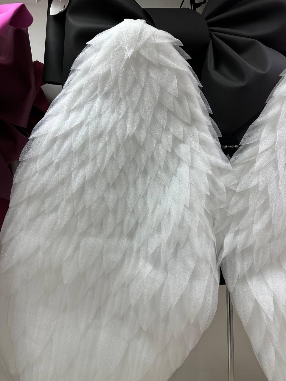 May include: Close-up of white, layered fabric wings with a feather-like design. The wings are made of a soft, textured material. A black bow is visible in the background, along with a dark pink bow.