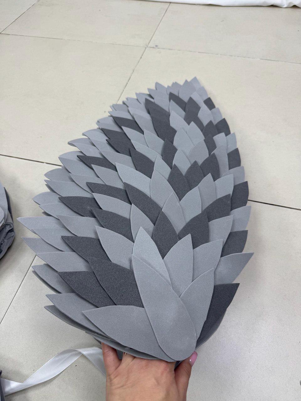 May include: A gray and dark gray felt feather-shaped accessory. The accessory is made of layered felt cut into leaf shapes. A white ribbon is attached to the base. The accessory is held by a hand.