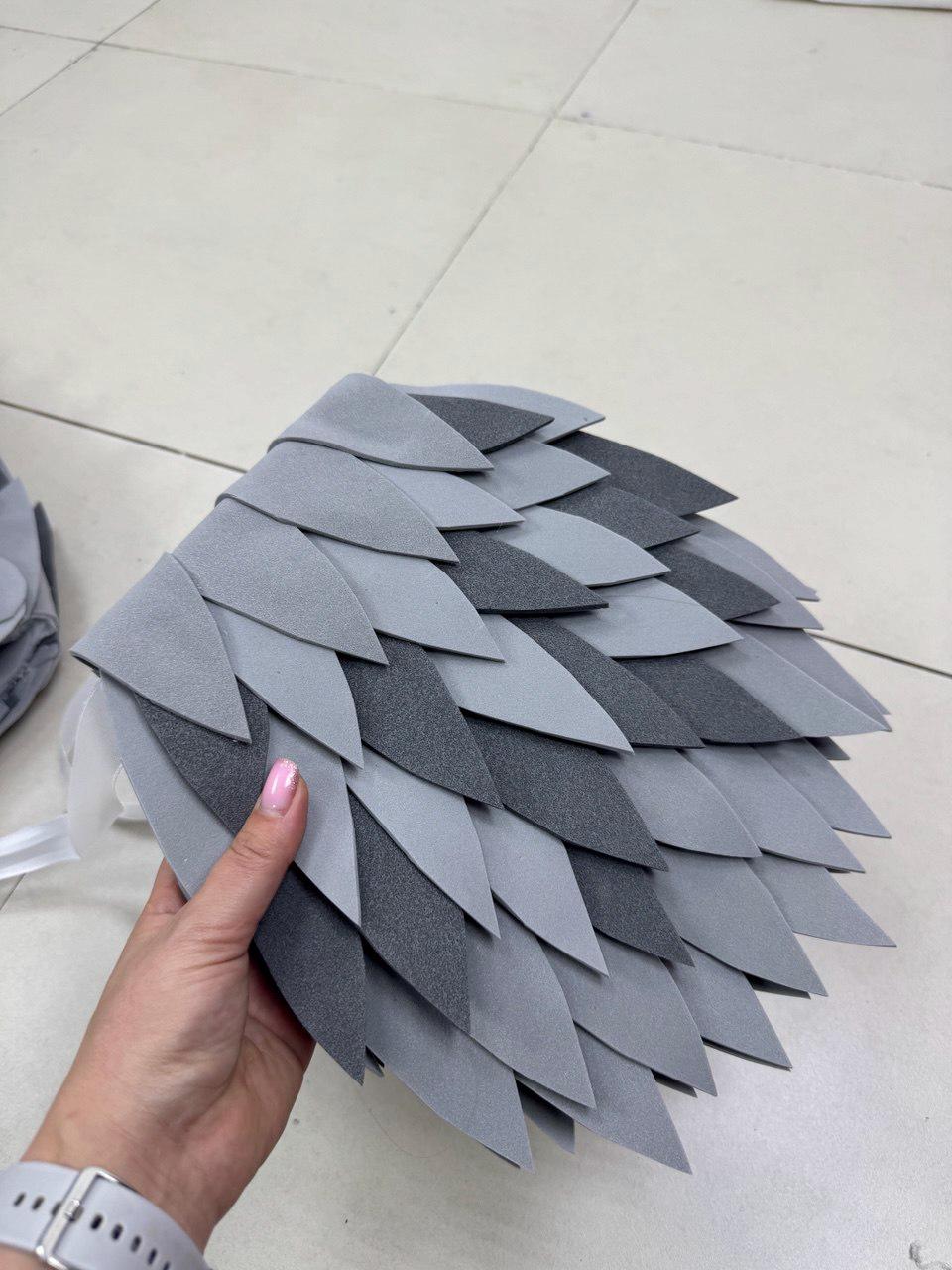 May include: A layered, gray wing prop with a feather-like design. The wing features various shades of gray, creating a textured appearance. The wing is held by a person, showcasing the size and detail of the costume accessory.