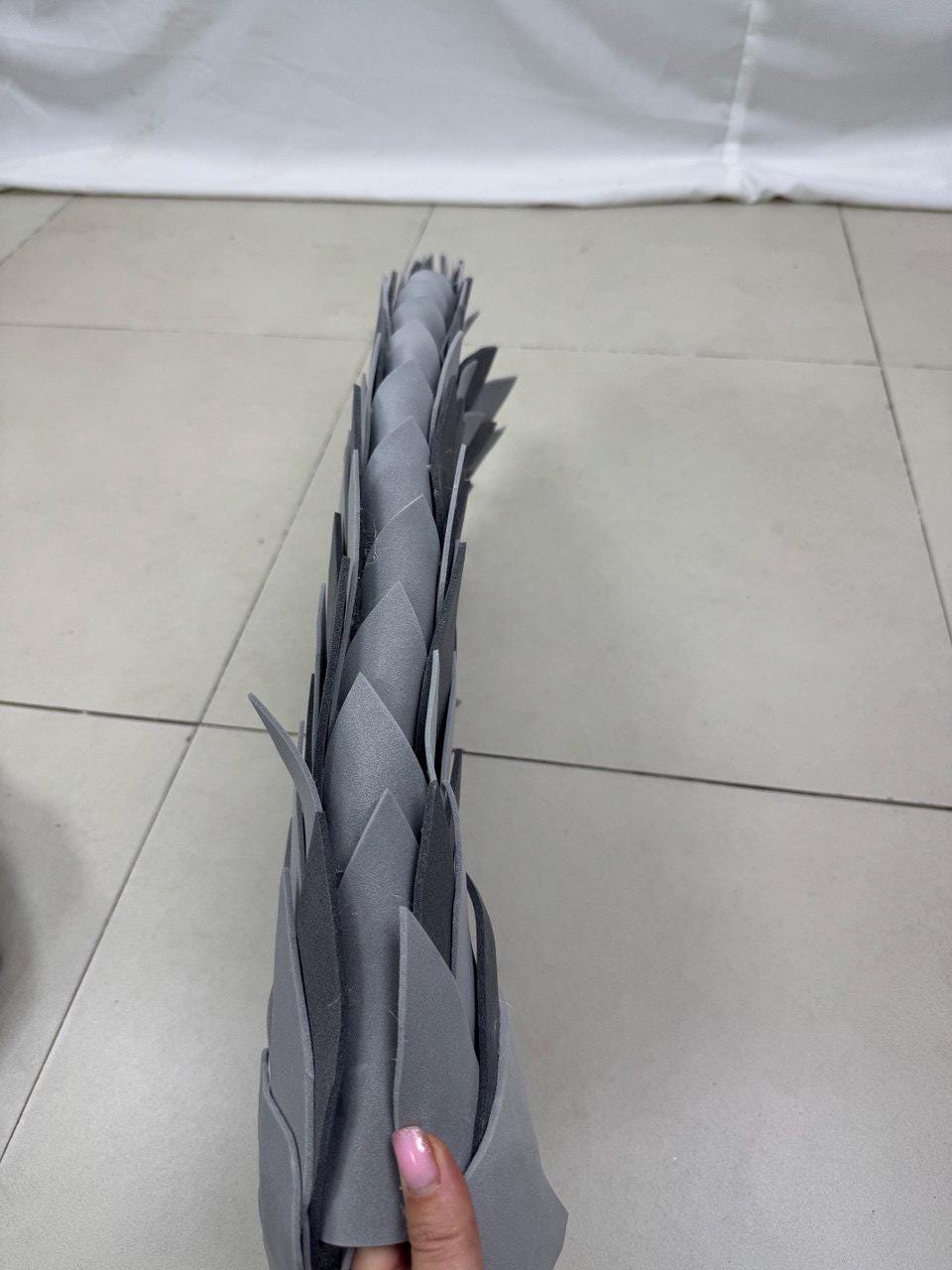 May include: A gray, layered, decorative object with a pointed, leaf-like design. The object is made of a soft material and has a textured appearance. The object is held by a person's hand.