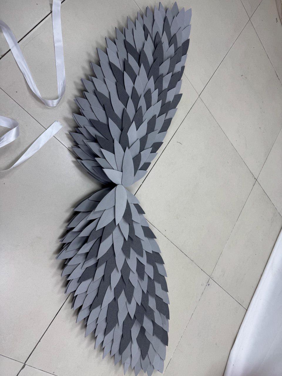 May include: A pair of gray and dark gray layered feather wings with white ribbon ties. The wings are made of foam and are arranged in a symmetrical pattern. The wings are designed for costume or cosplay use.