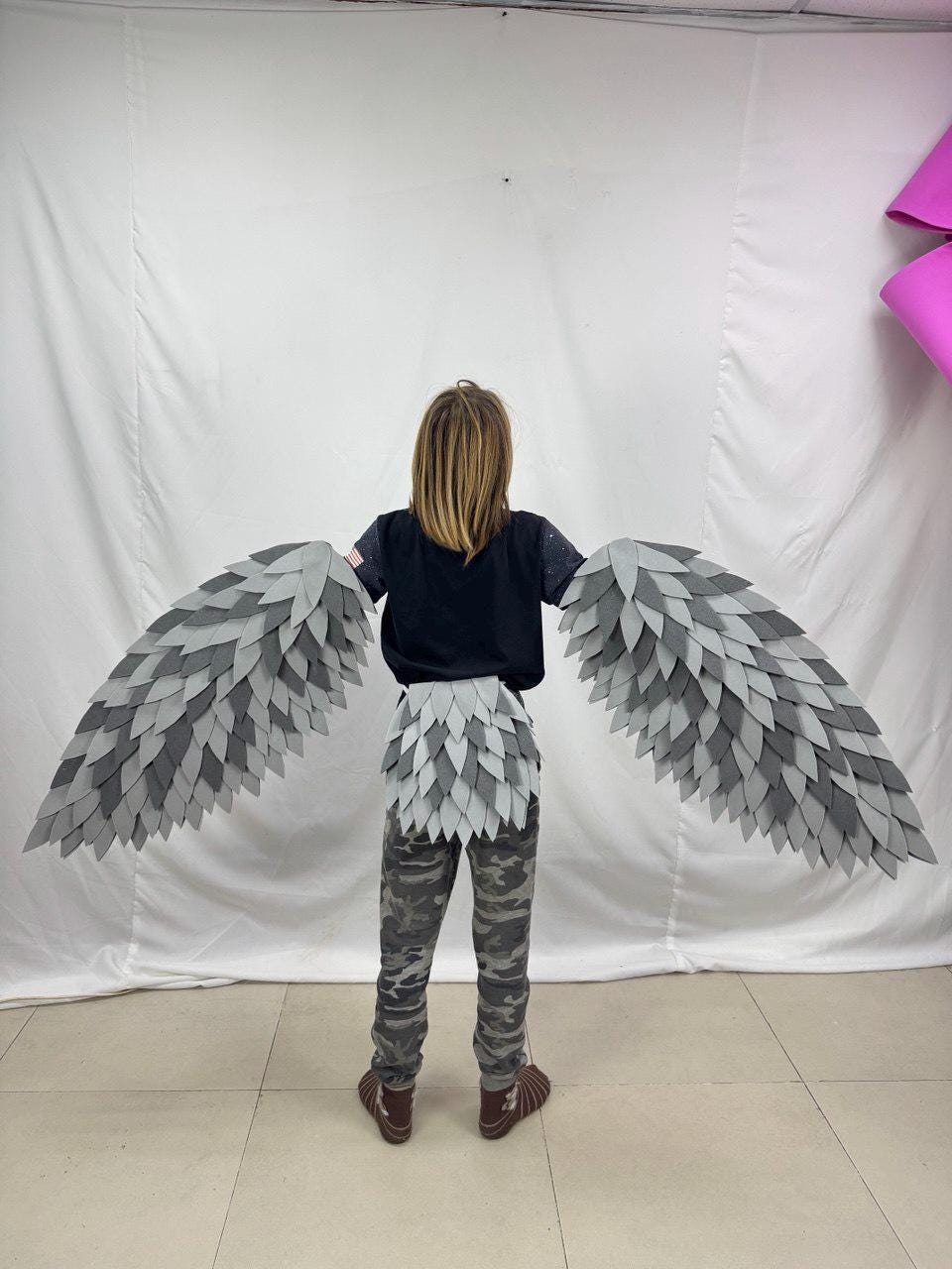May include: A costume featuring large, layered gray and white wings, and a matching waist piece. The wings extend outward, creating a dramatic effect. The costume is designed to resemble a bird or mythical creature. The person is wearing a black shirt and camouflage pants.