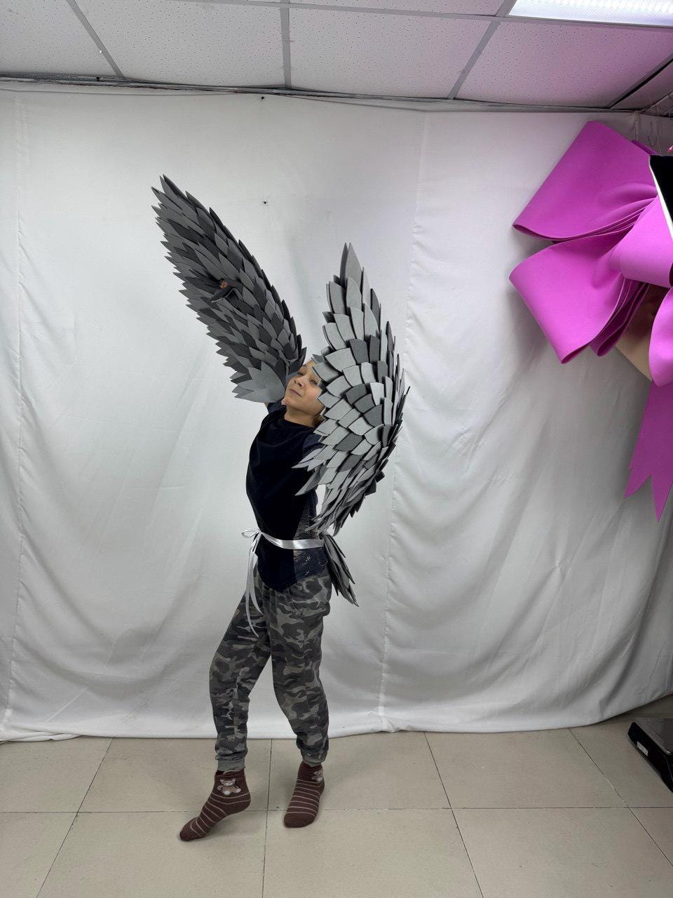 May include: A person wearing a set of gray and silver feathered wings. The wings are attached to the back with a white ribbon. The person is wearing a black shirt, camouflage pants, and brown socks. The wings are spread wide.
