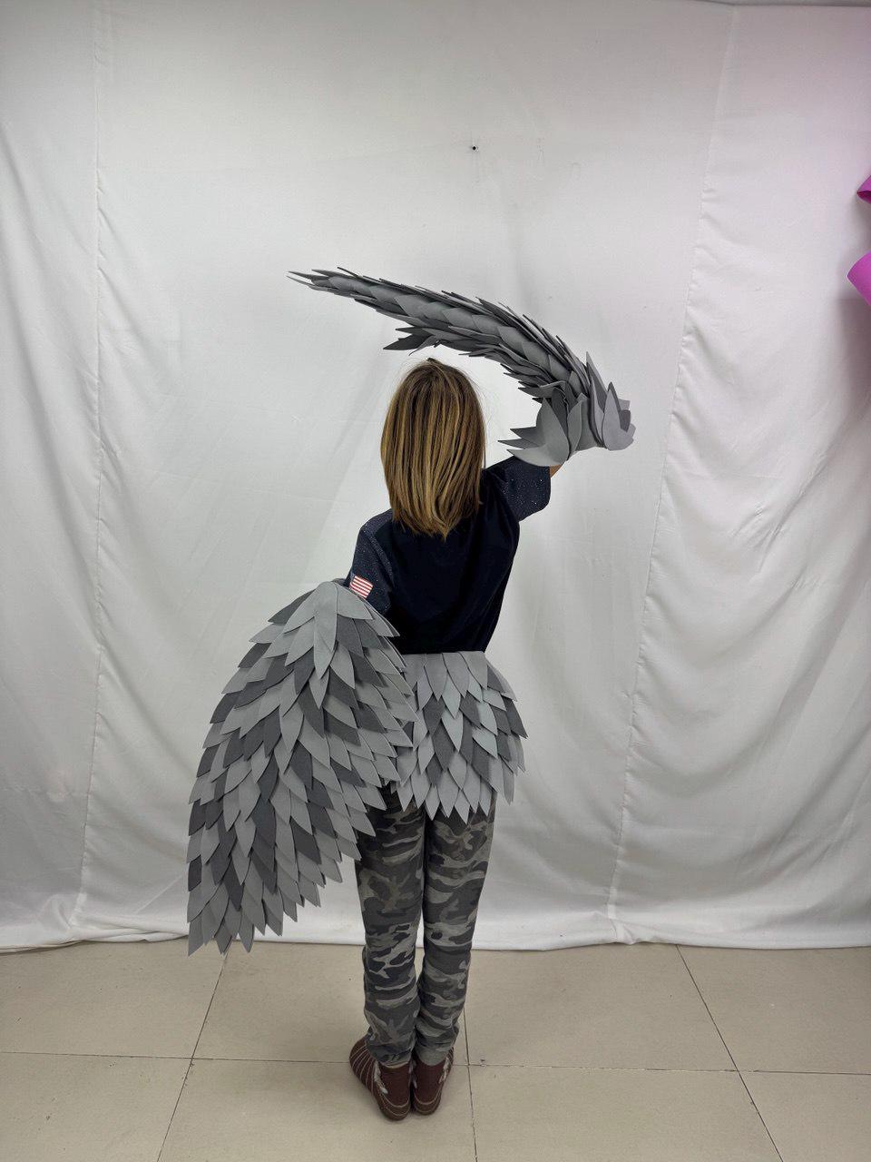 May include: A child wearing a gray and light gray feather-like costume, with matching wings and a skirt-like piece. The costume is made of layered, textured material. The child is wearing camouflage pants and a dark blue shirt.