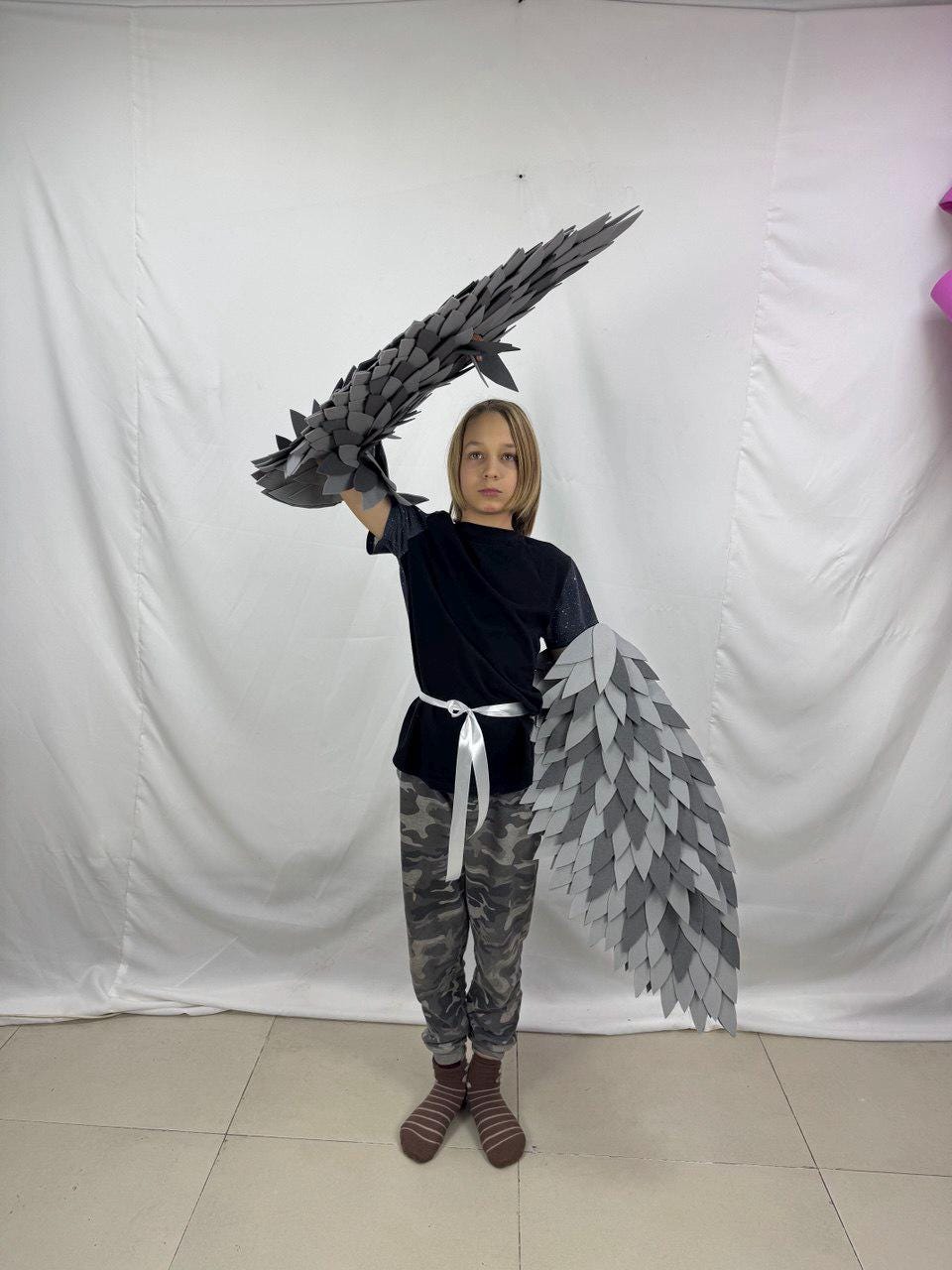 May include: A costume featuring gray feather-like wings and arm pieces. The wings are constructed from layered, textured gray material. The arm pieces are designed to mimic the appearance of feathers. The costume is designed to be worn.