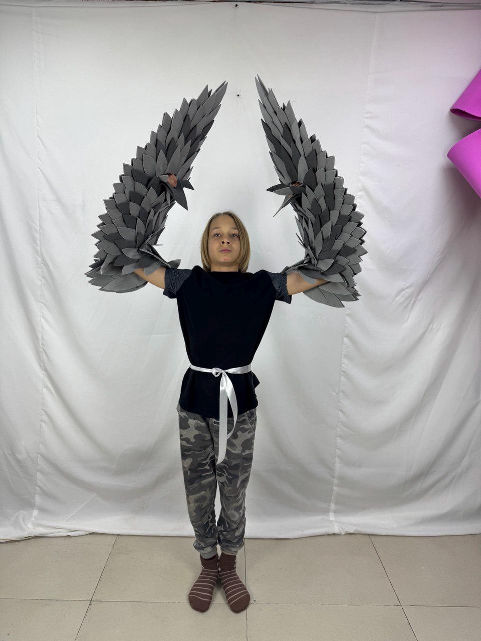 May include: A person wearing a black shirt, camouflage pants, and gray and white striped socks, holding up a pair of gray, layered, feather-like wings. The wings are made of multiple layers of gray material.