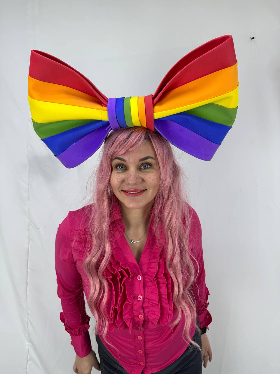 May include: A large, rainbow-colored bow headband. The bow features the colors of the Pride flag: red, orange, yellow, green, blue, and purple. The headband is worn by a person with pink hair and a pink shirt.