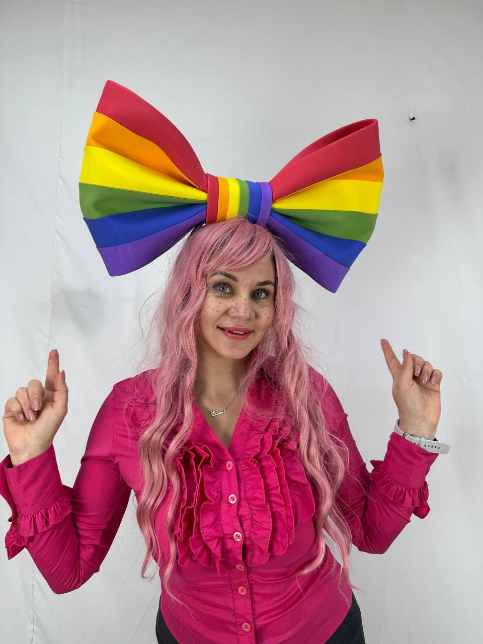 May include: A large rainbow-colored bow headband. The bow features the colors of the rainbow: red, orange, yellow, green, blue, and purple. The headband is worn by a person with pink hair and a pink shirt.