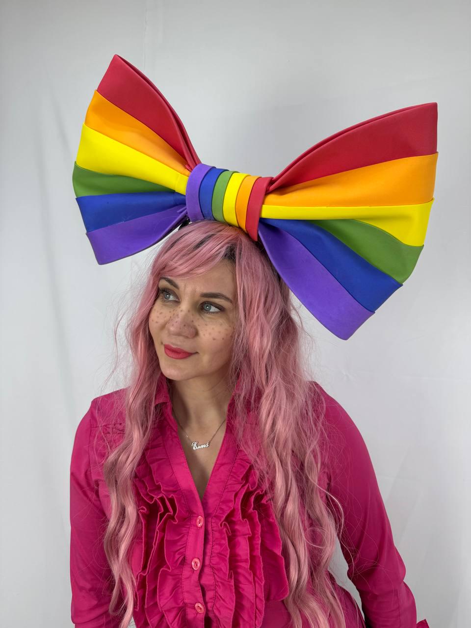 May include: A large, rainbow-colored bow headband. The bow features the colors of the rainbow: red, orange, yellow, green, blue, and purple. The headband is a festive accessory.