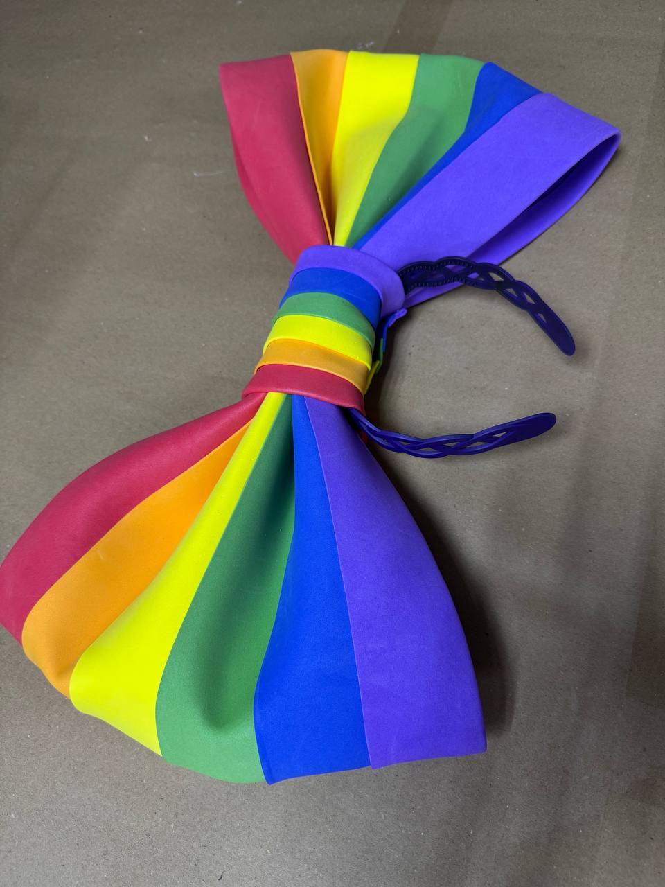 May include: A large, colorful rainbow bow tie. The bow tie features a spectrum of colors, including red, orange, yellow, green, blue, and purple. The bow tie is made of a soft material and has a dark blue adjustable strap.