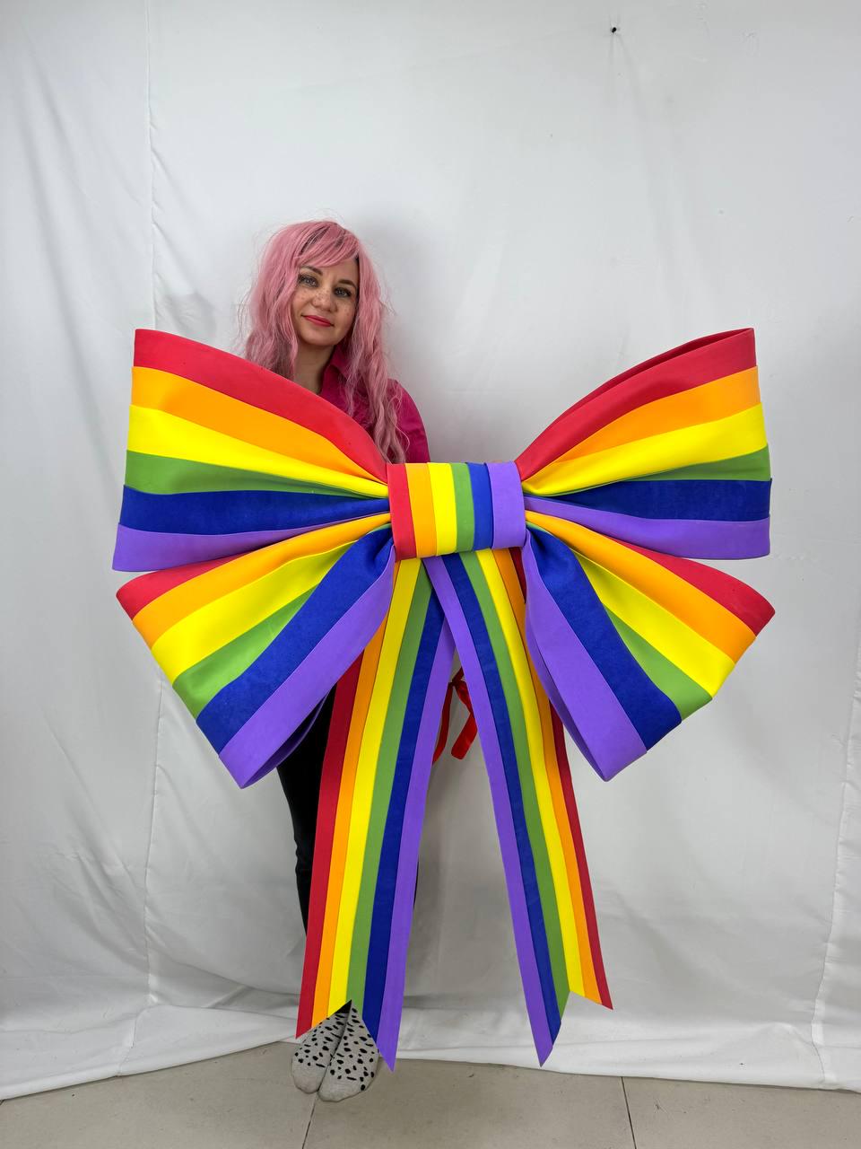 May include: A large, colorful rainbow bow with red, orange, yellow, green, blue, and purple stripes. The bow is held up against a white backdrop. The bow's dimensions are approximately 48 inches wide and 36 inches tall.