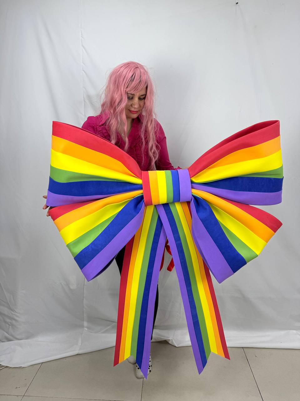 May include: A large, colorful rainbow bow with red, orange, yellow, green, blue, and purple stripes. The bow is held by a person wearing a pink shirt. The bow's design is a symbol of LGBTQ+ pride.