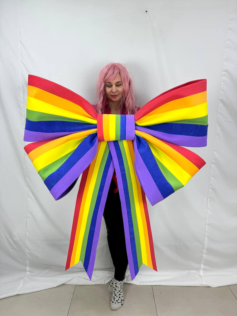 May include: A large, colorful rainbow bow with red, orange, yellow, green, blue, and purple stripes. The bow is worn by a person wearing black pants and a pink shirt. The bow's streamers hang down.