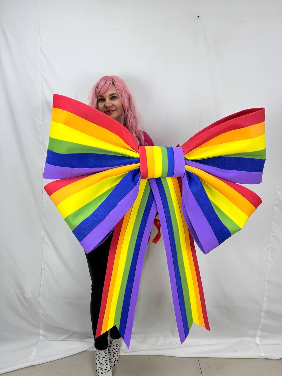 May include: A large, colorful rainbow bow with red, orange, yellow, green, blue, and purple stripes. The bow is a vibrant decoration, perfect for a party or event. The bow is held by a person.