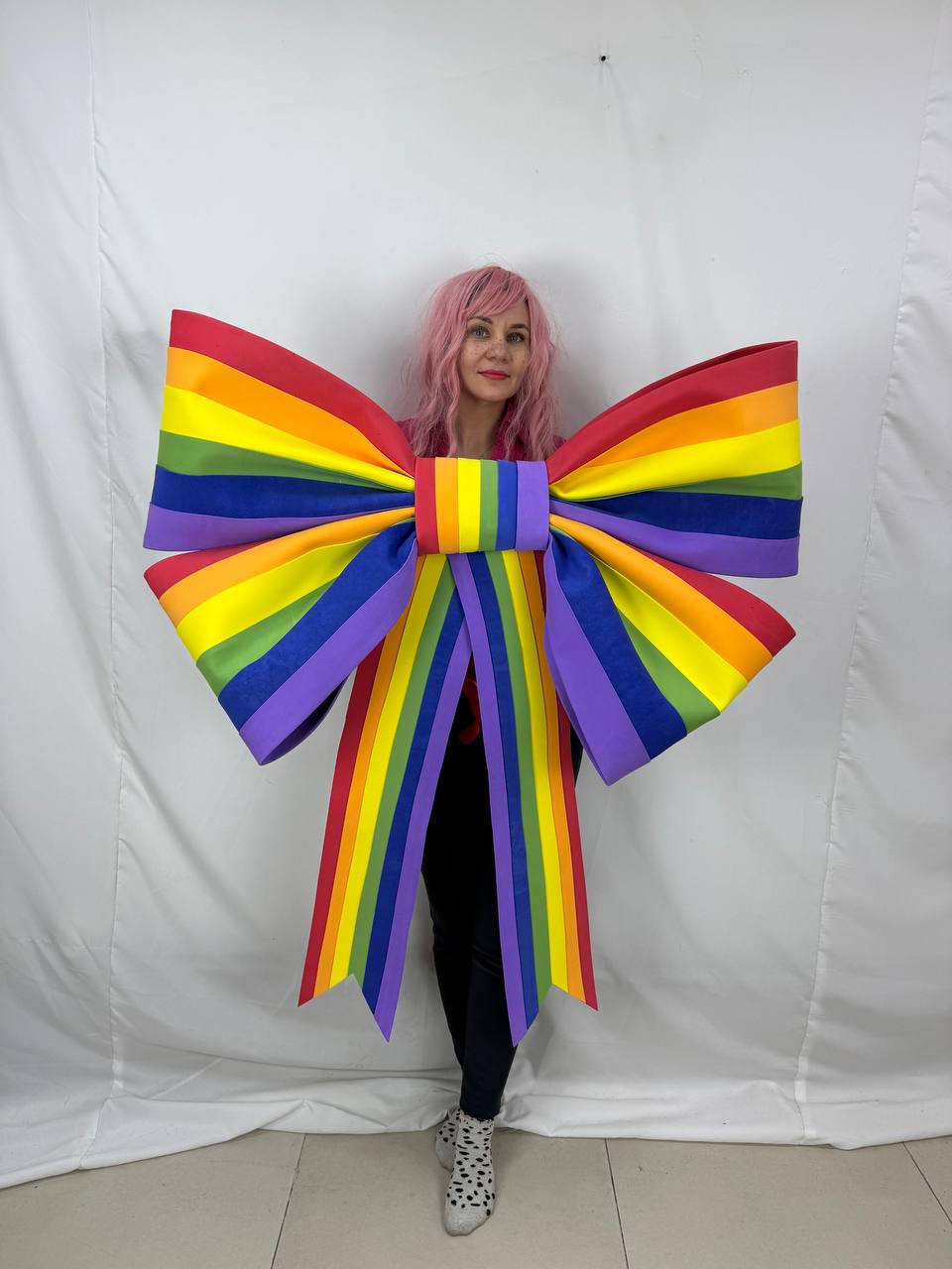 May include: A large, colorful rainbow bow costume with red, orange, yellow, green, blue, and purple stripes. The bow is worn by a person with pink hair and black pants. The bow's ribbons hang down.