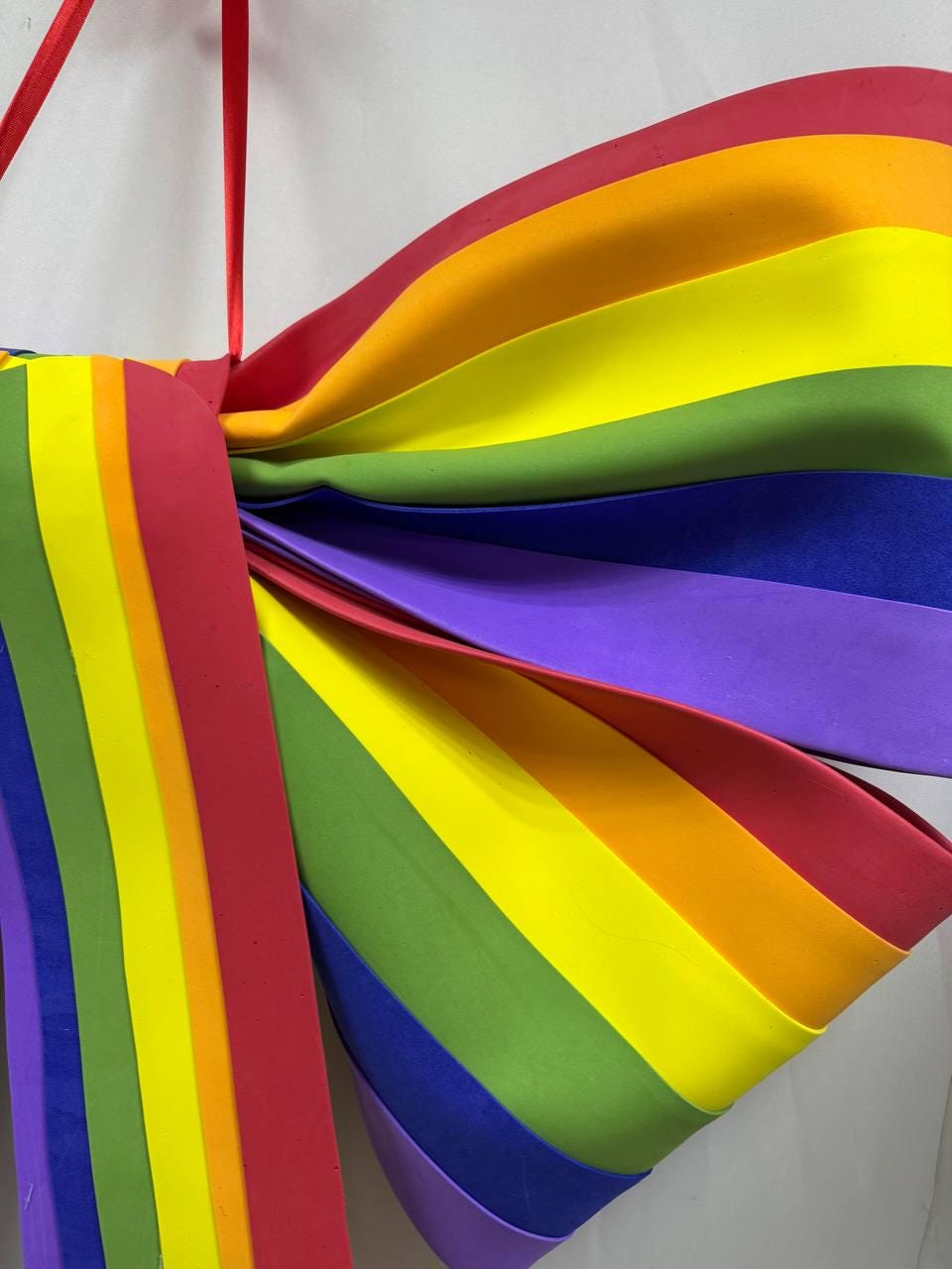 May include: A large, colorful rainbow-themed decoration. The design features layered, curved sections in red, orange, yellow, green, blue, and purple. A red ribbon is attached at the top for hanging. The overall shape resembles a large bow or butterfly wings.