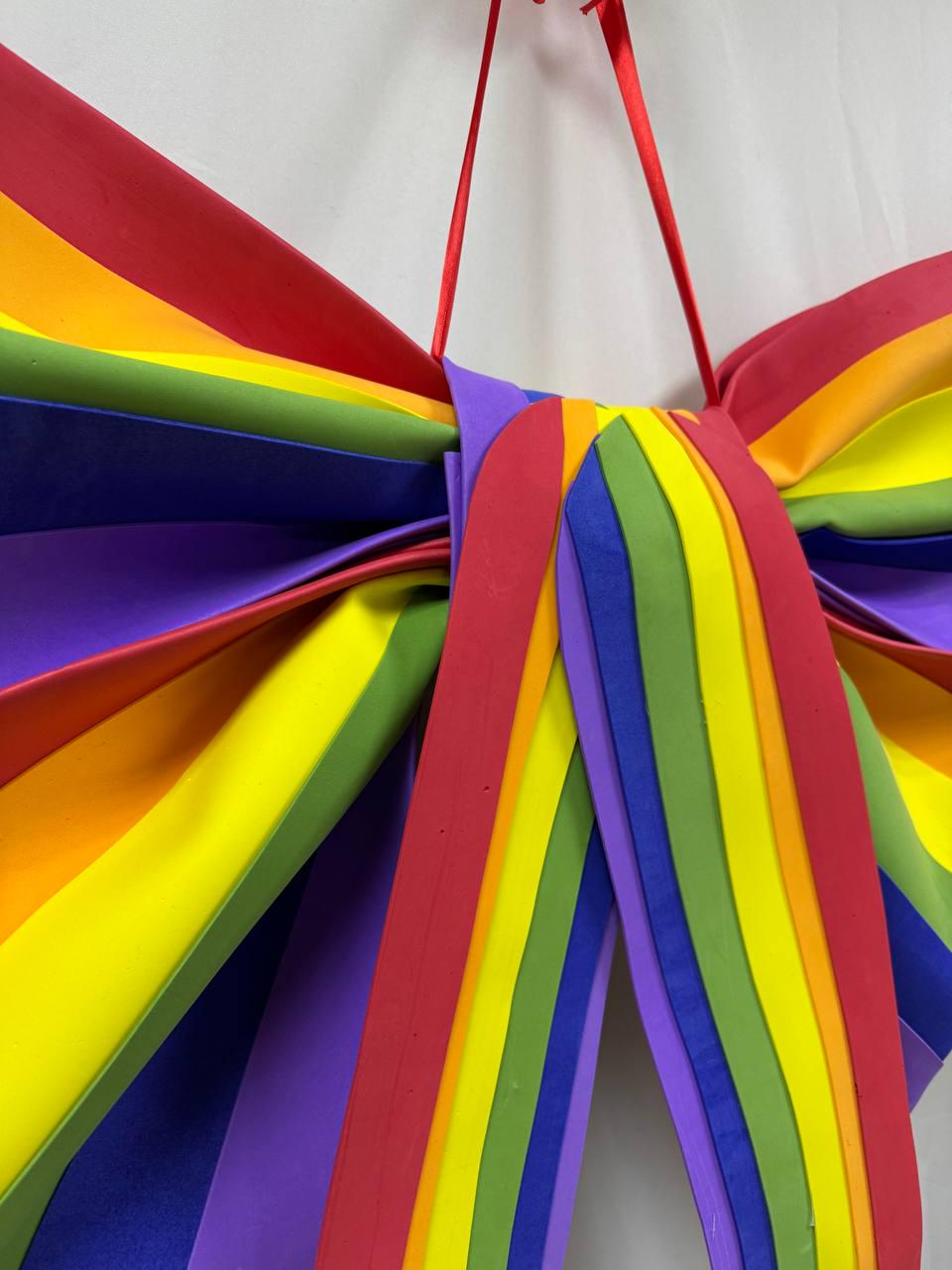May include: A large, colorful rainbow bow with red, orange, yellow, green, blue, and purple stripes. The bow is made of layered material and has a red ribbon for hanging.