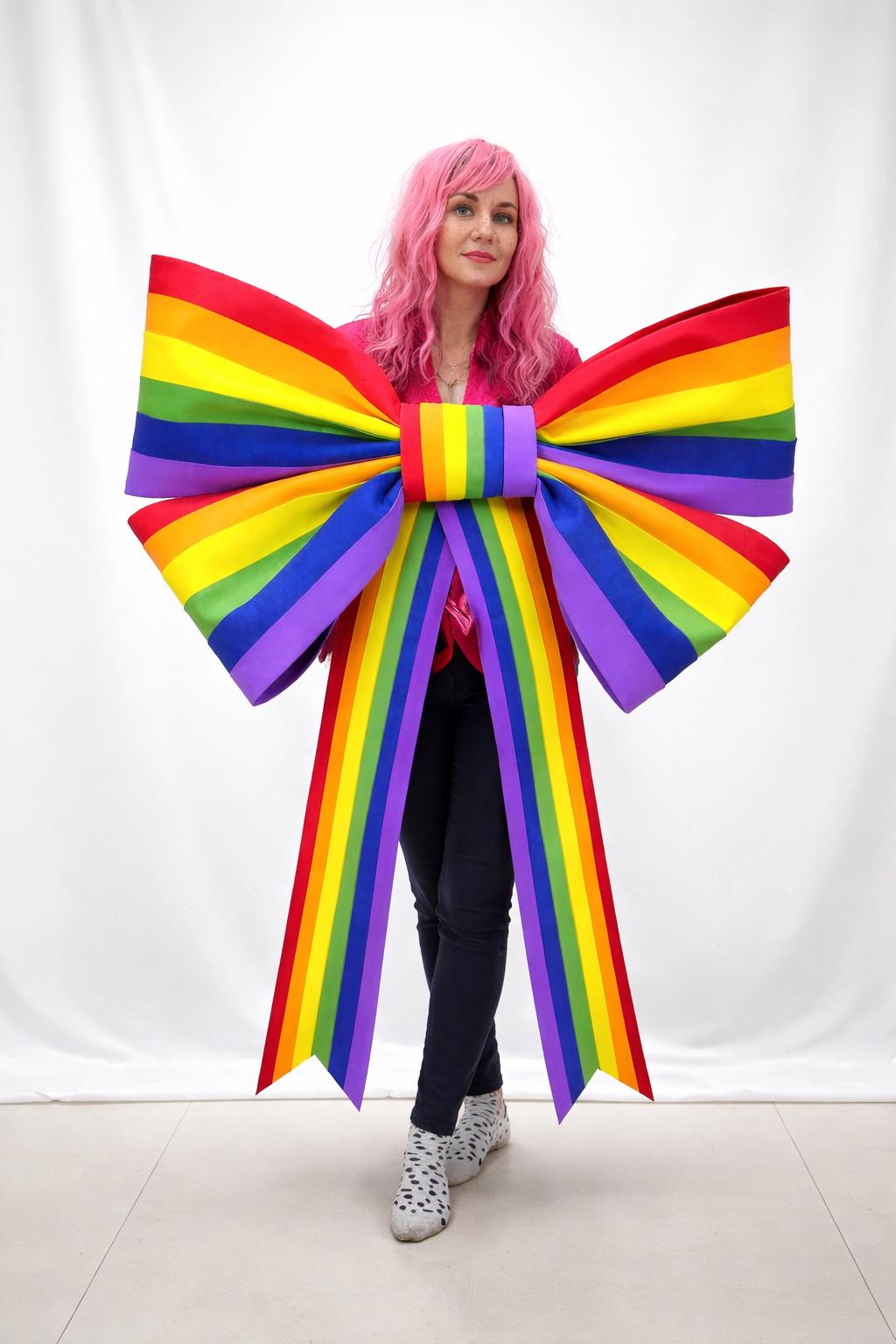 May include: A large, colorful rainbow bow with red, orange, yellow, green, blue, and purple stripes. The bow is held in front of a person wearing a pink shirt and black pants. The bow's ribbons extend downwards.