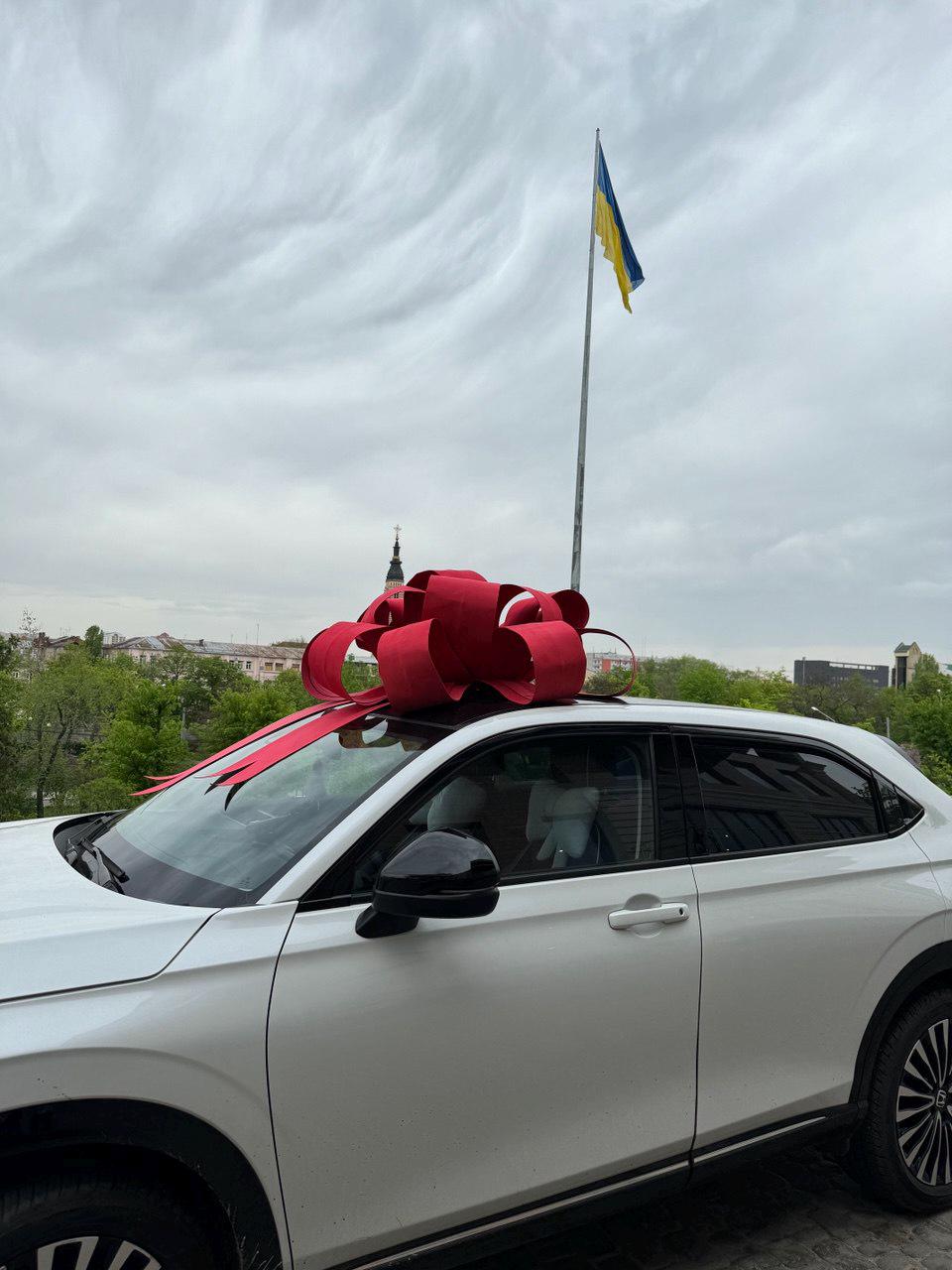 May include: A white car with a large red bow on the roof. The car is parked outside with a Ukrainian flag waving in the background. The sky is overcast.