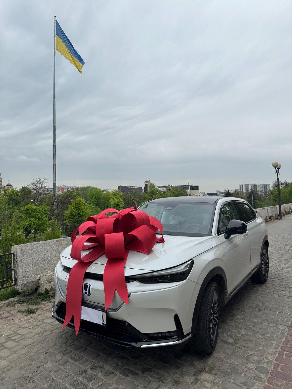 May include: A white SUV with a large red bow on the hood. The car is parked on a brick-paved road with a Ukrainian flag waving in the background. The sky is overcast.