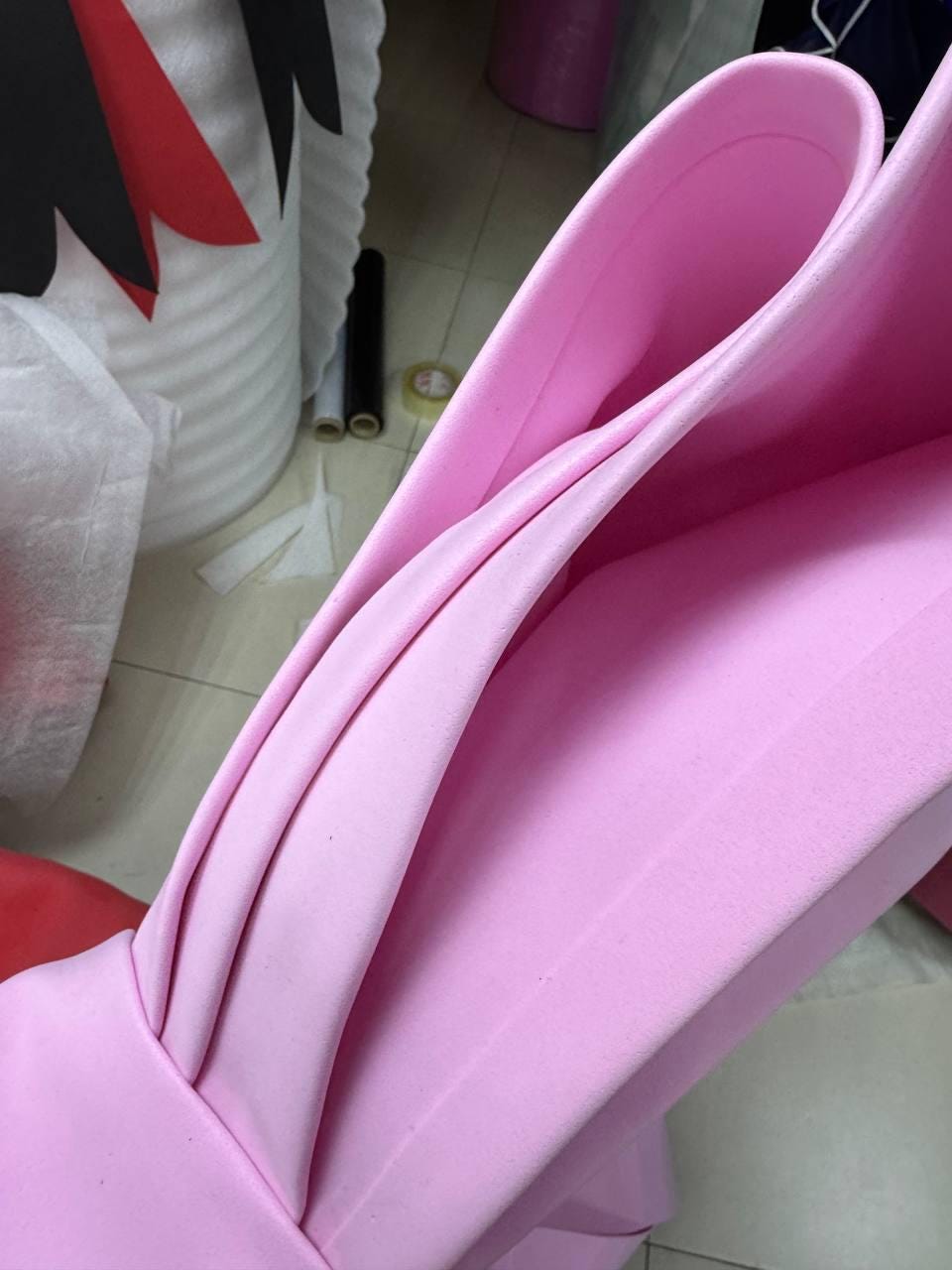 May include: Close-up of a large, pink, curved foam structure. The foam has a smooth texture and is folded in a way that creates a layered effect. The edges are rounded, and the overall shape is reminiscent of a large bow or petal. The background shows other crafting materials.