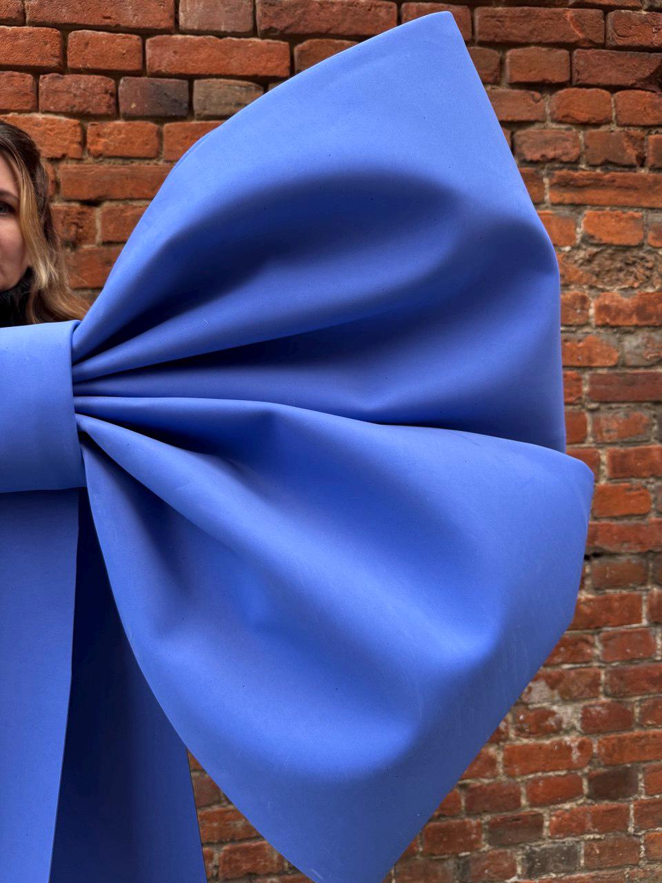 May include: A large, vibrant blue bow, likely for decoration or a gift. The bow is made of a smooth, matte material and is set against a rustic brick wall background. The bow's size suggests it's a statement piece.