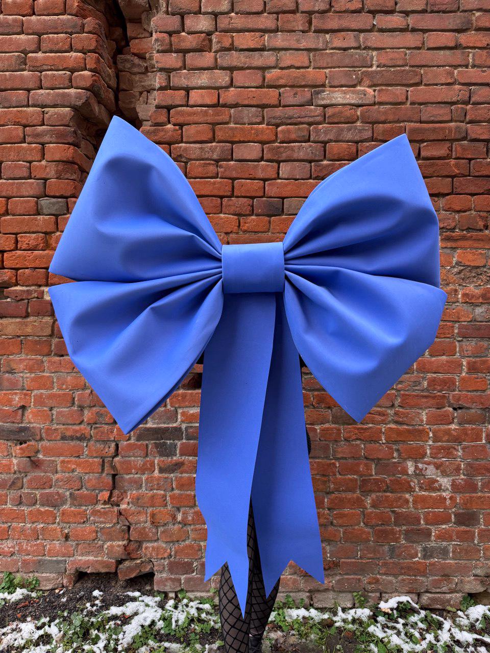 May include: A large, vibrant blue bow with long streamers. The bow is the focal point, set against a weathered red brick wall. The bow's size and color create a striking visual contrast.