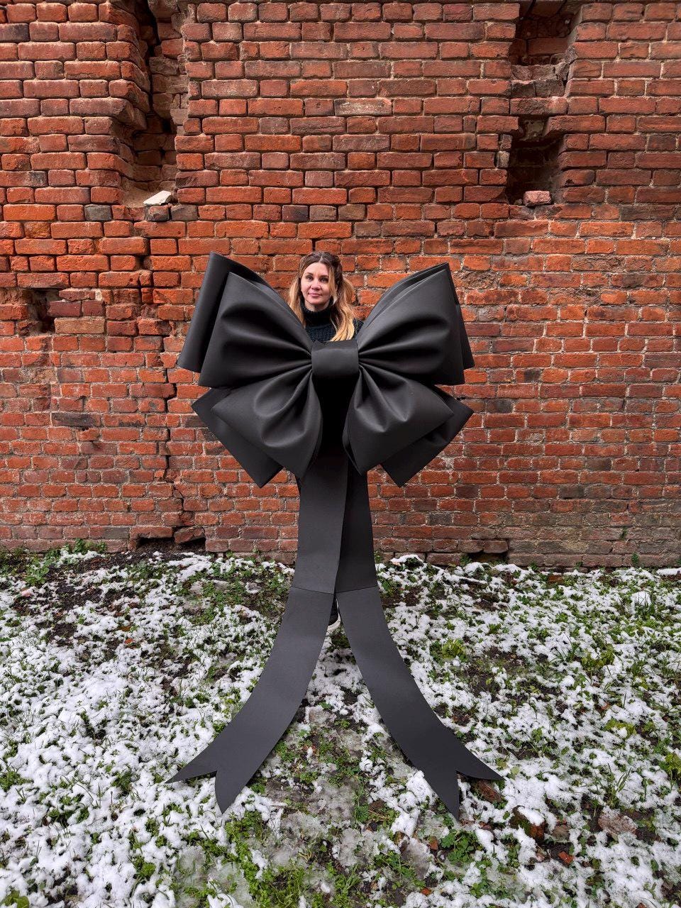 May include: A large, black bow with long, flowing tails. The bow is positioned in front of a red brick wall and a patch of snow-covered ground. The bow's size suggests it could be used for a large gift or decoration.