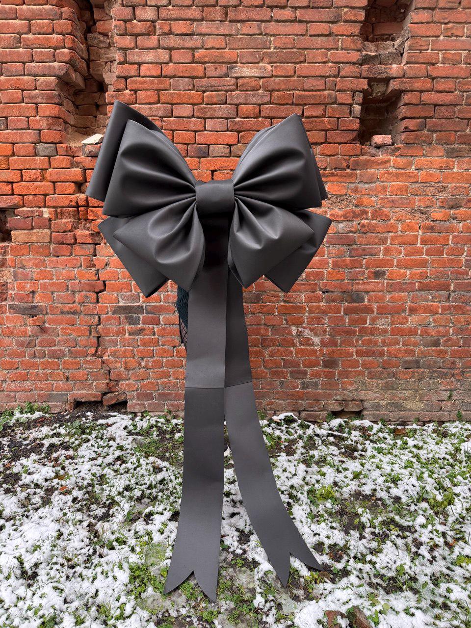 May include: A large, dark gray bow with long, pointed streamers. The bow is set against a weathered brick wall and rests on a patch of grass and snow. The bow's design is symmetrical and decorative.