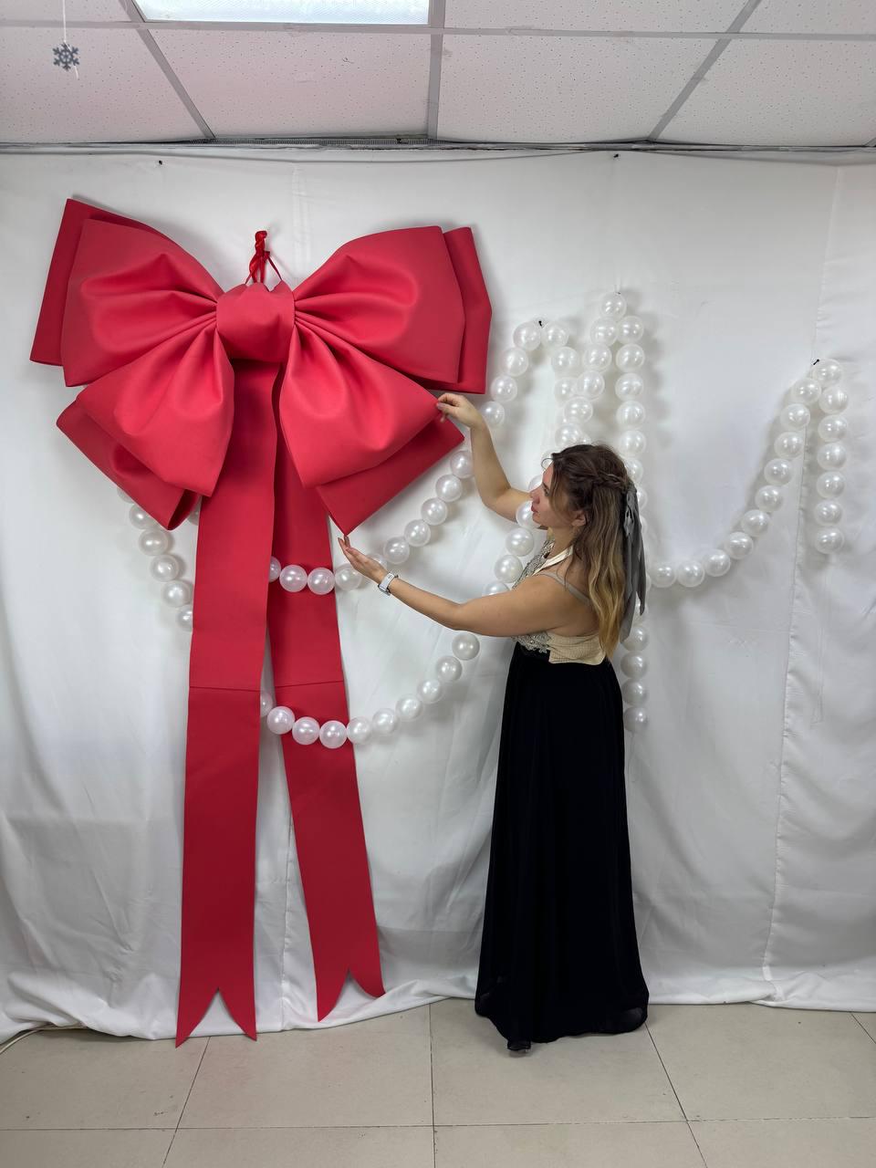 May include: A large, vibrant red bow with long, flowing ribbons, suspended against a white backdrop. The bow is adorned with a string of white pearls. The bow is a festive decoration.
