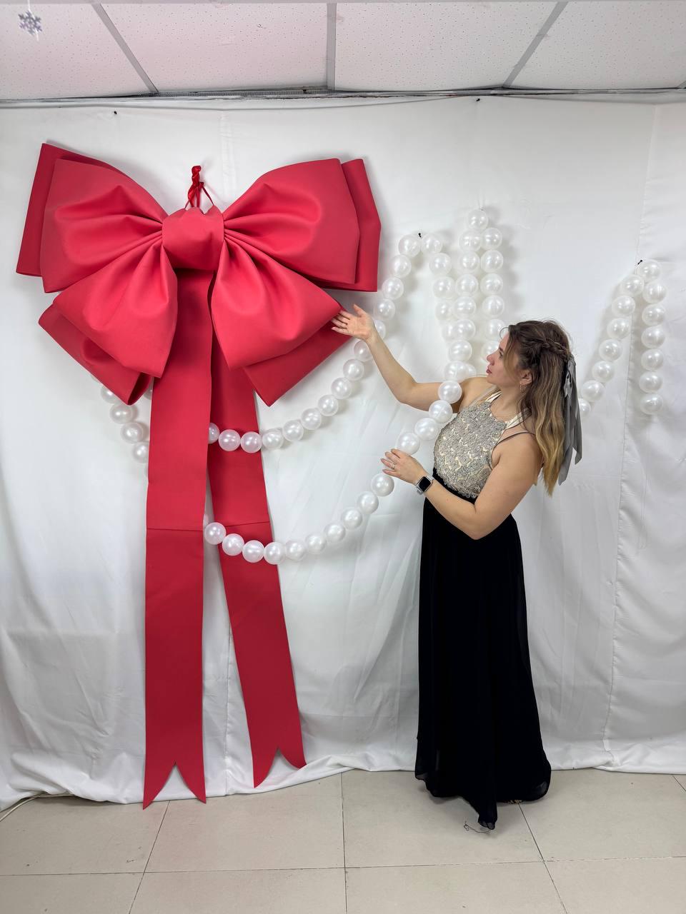 May include: A large, vibrant red bow with long streamers hangs against a white backdrop. A strand of white pearls is draped from the bow. The bow is a festive decoration, possibly for a holiday or special event.