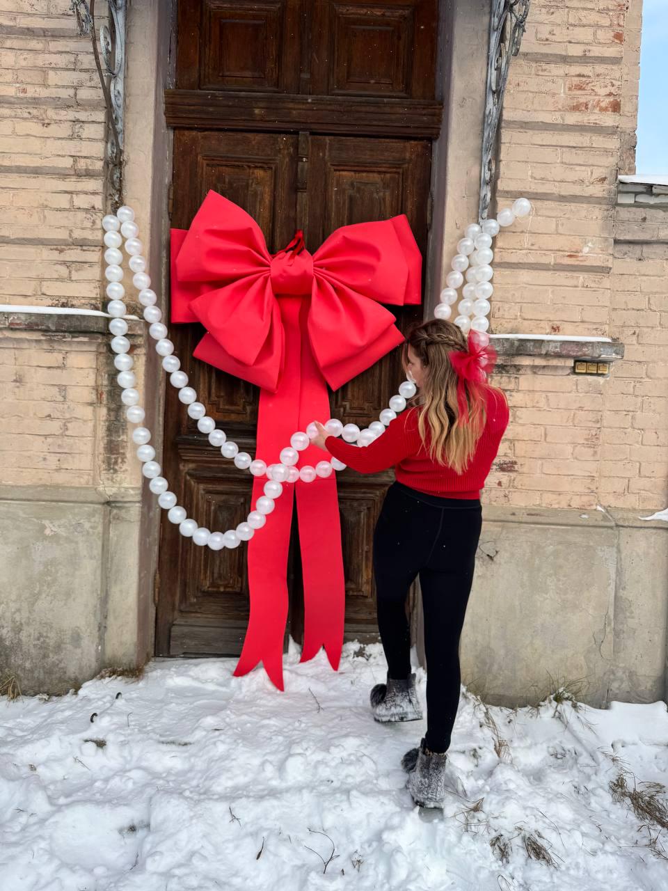 May include: A large, bright red bow adorns a wooden door, with long, flowing ribbons. White, spherical decorations hang on either side of the door. The scene is set outdoors, with snow on the ground, suggesting a festive holiday theme.
