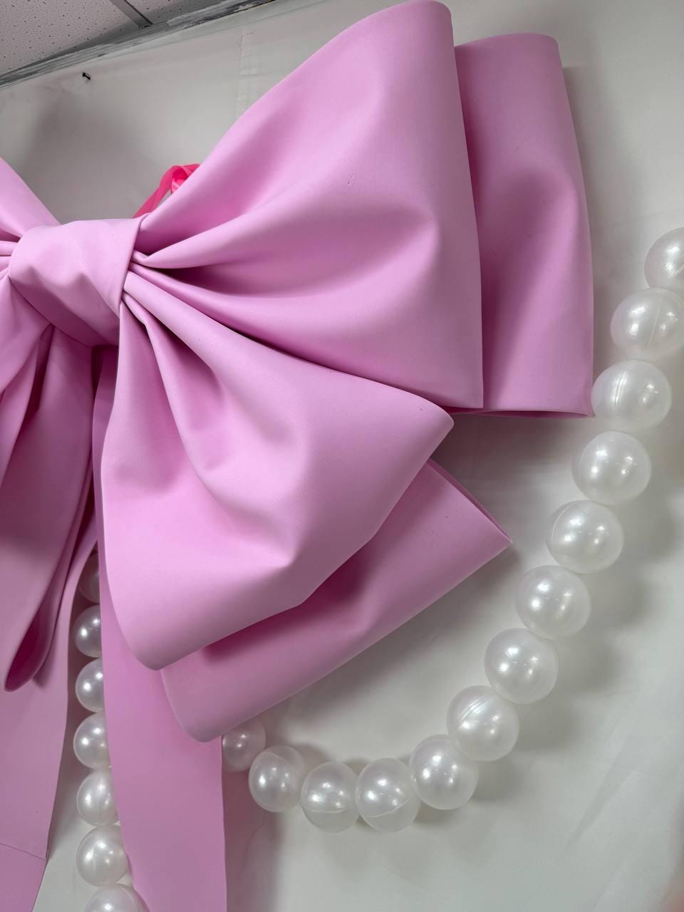 May include: A large, light pink bow with multiple layers and a satin finish. A strand of large, white, pearl-like beads hangs alongside the bow. The bow is a decorative element, possibly for a party or event.