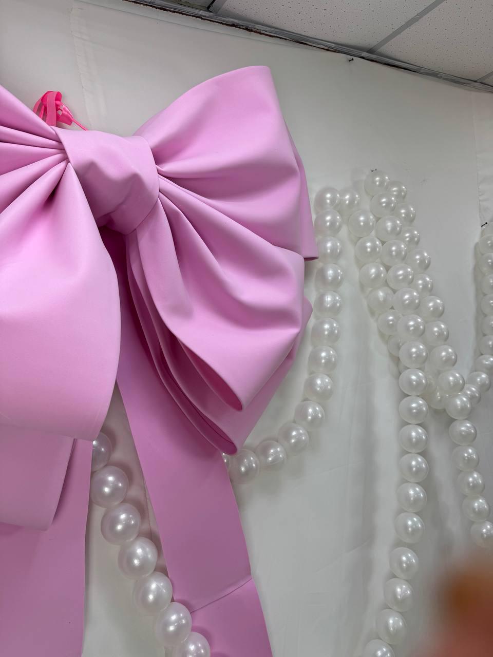 May include: A large, light pink bow with long, flowing ribbons is the focal point of this image. The bow is made of a smooth, satin-like material. Strands of white pearls are draped nearby, creating a decorative contrast against the white background.