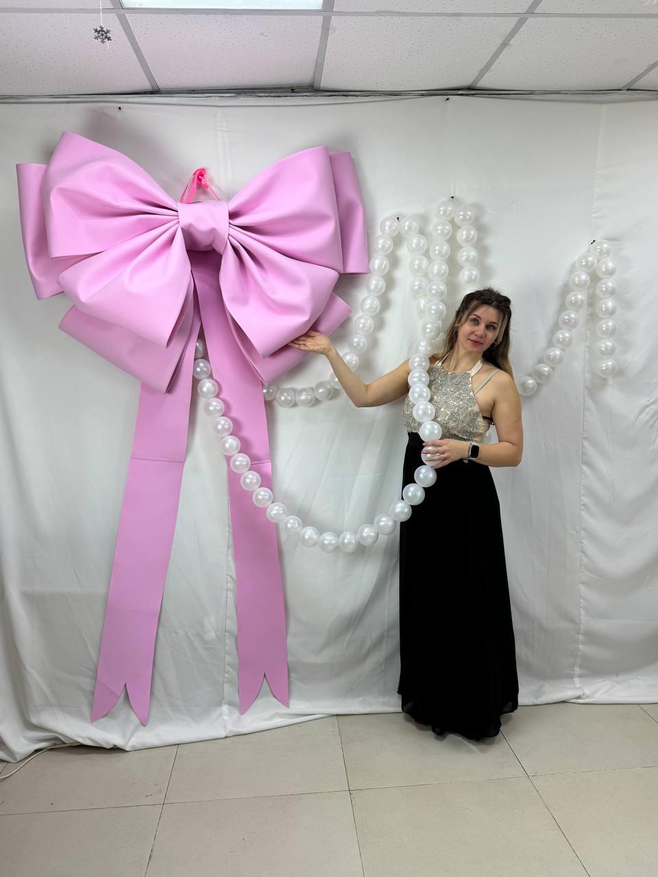 May include: A large, light pink bow with long streamers hangs against a white backdrop. A strand of large, white pearls drapes down from the bow. The bow is a decorative element, possibly for a party or event.