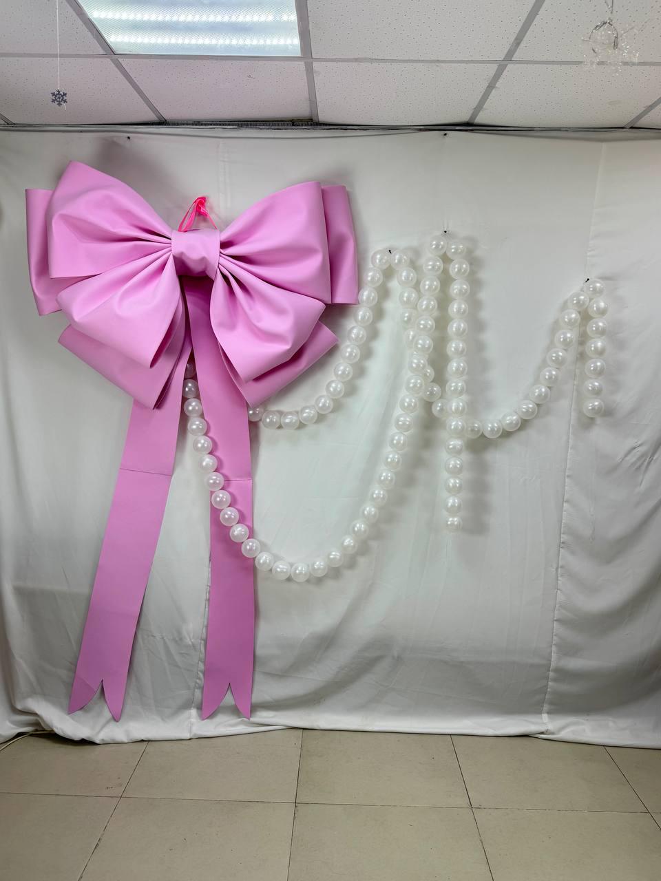 May include: A large, pink bow with long, pointed streamers hangs on a white backdrop. A strand of white pearls is draped to the right of the bow. The bow is a decorative element, possibly for a party or event.