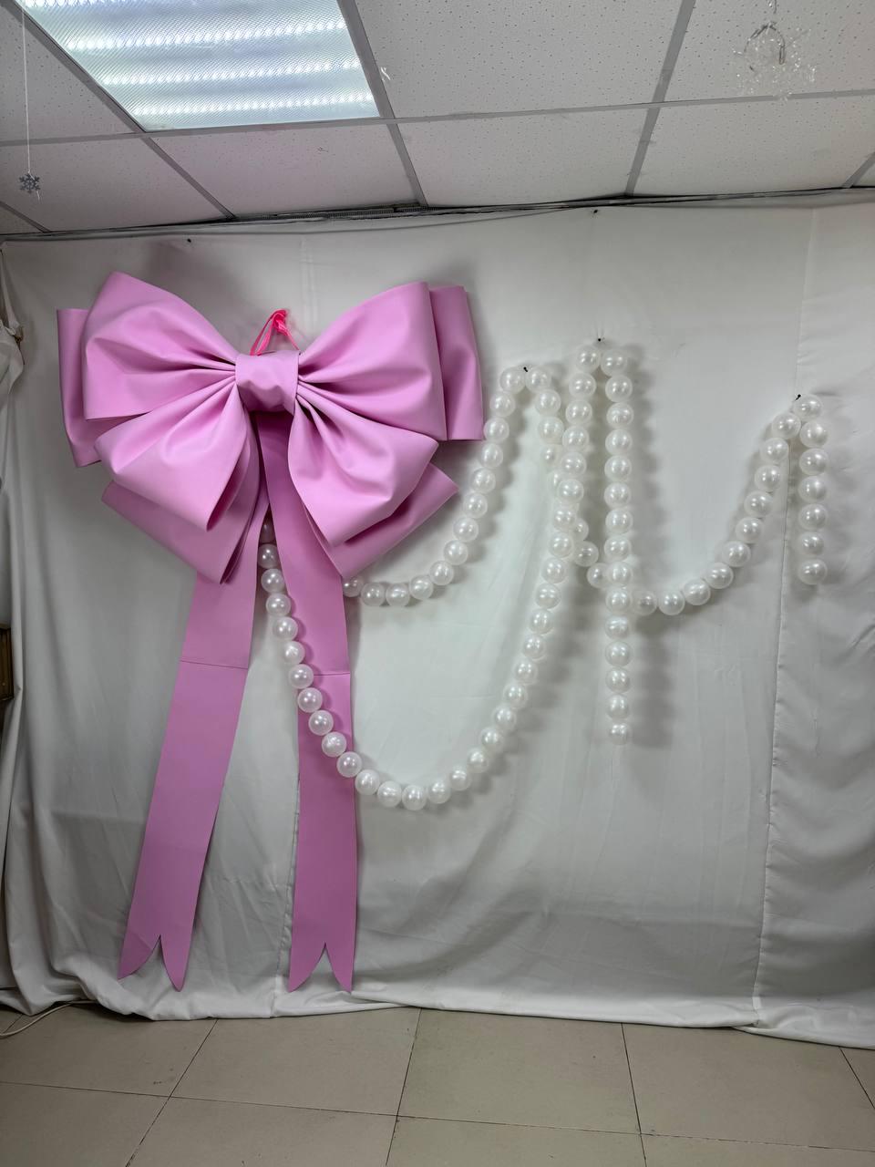 May include: A large, pink bow with long streamers hangs on a white backdrop. A strand of white, round beads is draped to the right of the bow. The bow is made of a smooth, possibly satin-like material, and is a vibrant shade of pink.