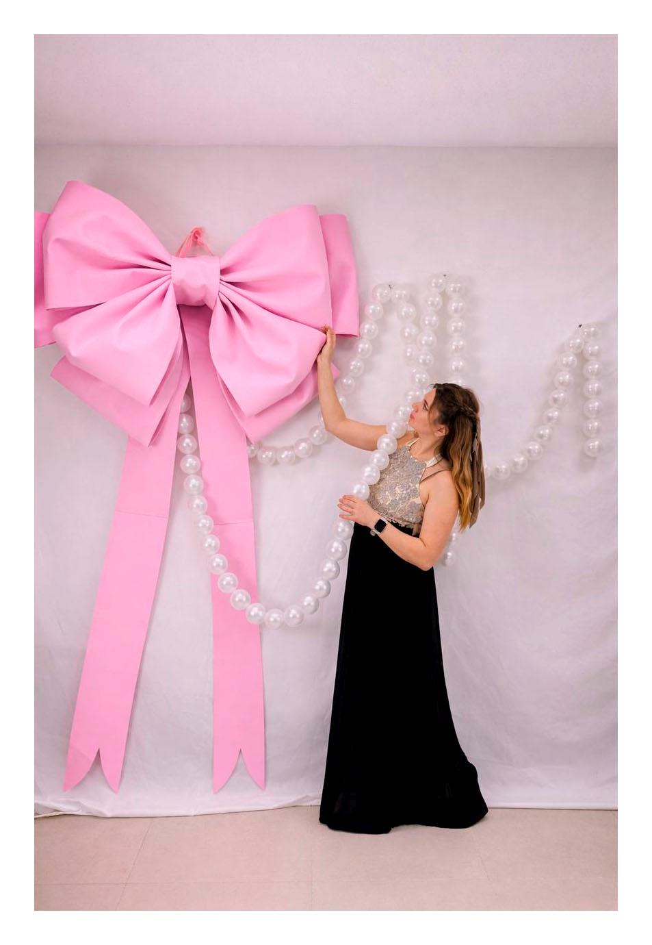 May include: A large, pink bow with long streamers is the focal point. A woman in a formal gown stands near the bow, holding a strand of pearl-like beads. The background is a plain white wall.