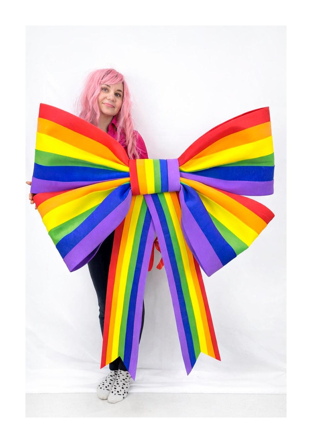 May include: A large, colorful rainbow bow with red, orange, yellow, green, blue, and purple stripes. The bow is held by a person with pink hair, wearing a pink shirt and black pants. The bow is a decorative item.