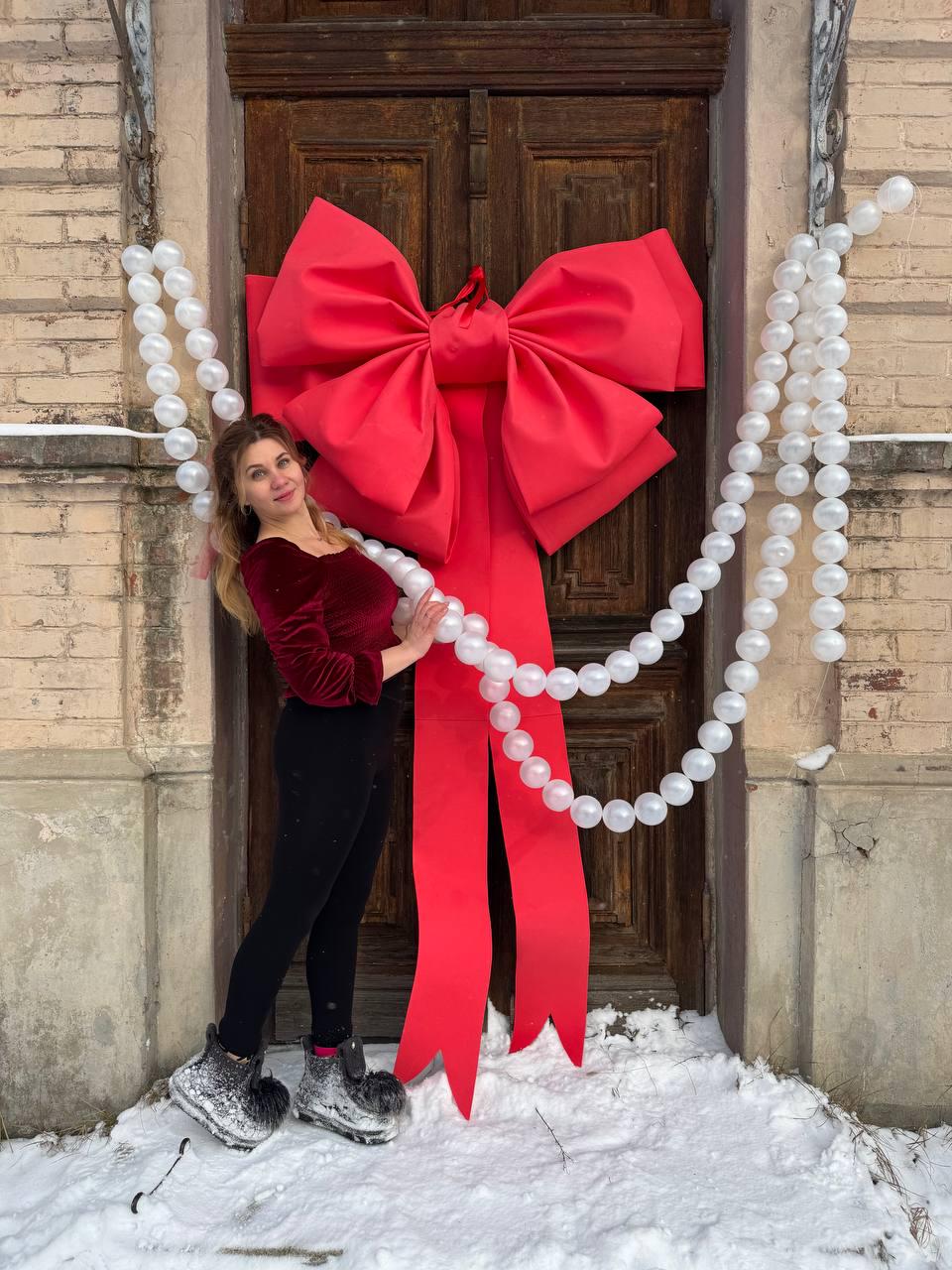 May include: A large red bow and matching ribbon adorn a wooden door, complemented by strings of white, round ornaments. The scene is set outdoors, with snow on the ground. The bow is the focal point of the festive decoration.