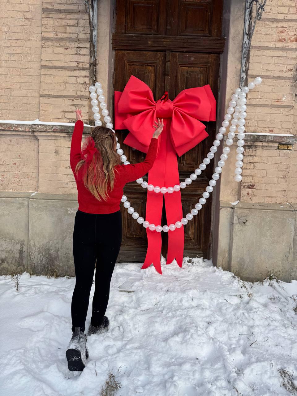 May include: A large red bow adorns a wooden door, with white beaded garlands draped on either side. A person wearing a red sweater and black pants stands in front of the door, arms raised. Snow covers the ground.