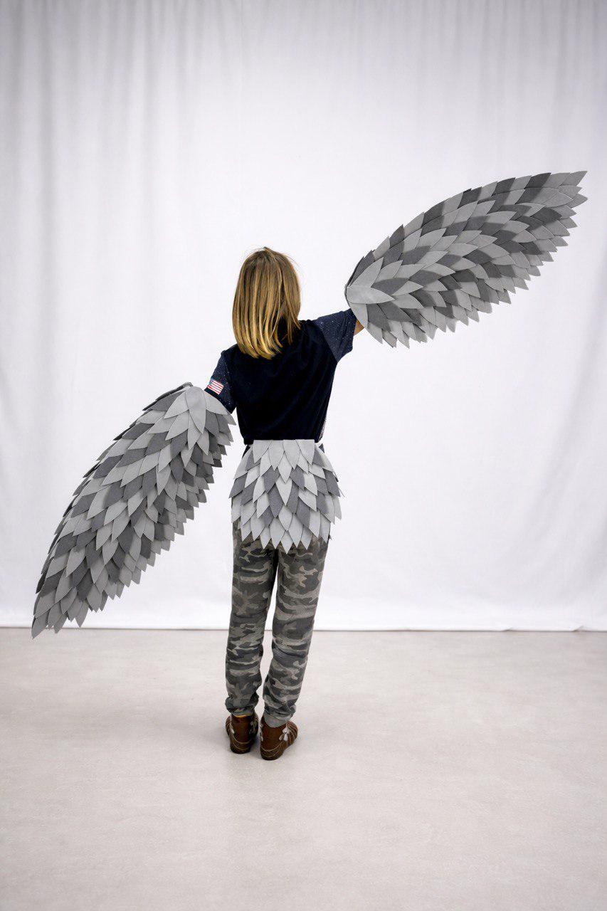 May include: A child wearing a gray and white feathered wing costume with matching waist piece. The wings are outstretched, showcasing the layered design. The child is wearing a dark blue shirt and camouflage pants.