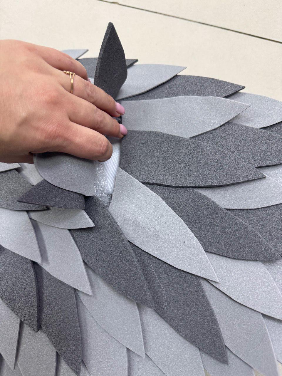 May include: A close-up of a layered, gray and silver decorative piece. The piece is composed of overlapping, leaf-shaped elements in varying shades of gray, creating a textured, dimensional effect. A hand with a gold ring is touching the piece.